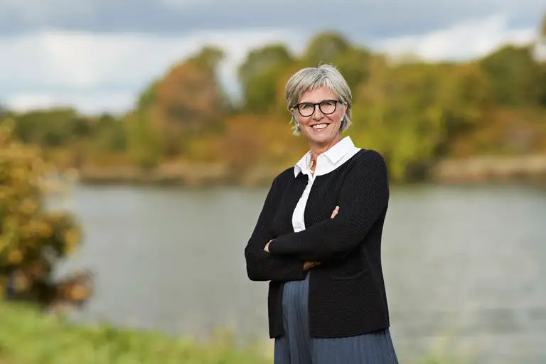 Bettina Lundgren i hvid skjorte, sort cardigan, blå nederdel. Bettina står med siden lidt til, armene over kors og smil med tænder. Billedet er beskåret lidt over knæet.