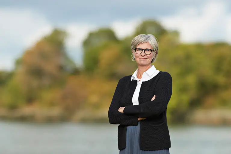 Bettina Lundgren i hvid skjorte, sort cardigan og blå nederdel. Hun står lidt skråt med armene over kors. Billedet er beskåret lidt over knæet.